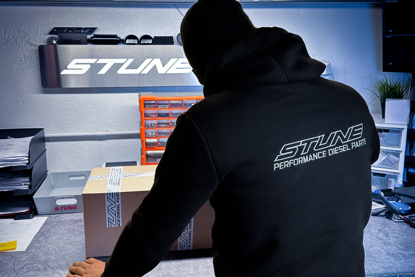 Person wearing a black hoodie with 'Stune Performance Diesel Parts' logo in a workshop setting.